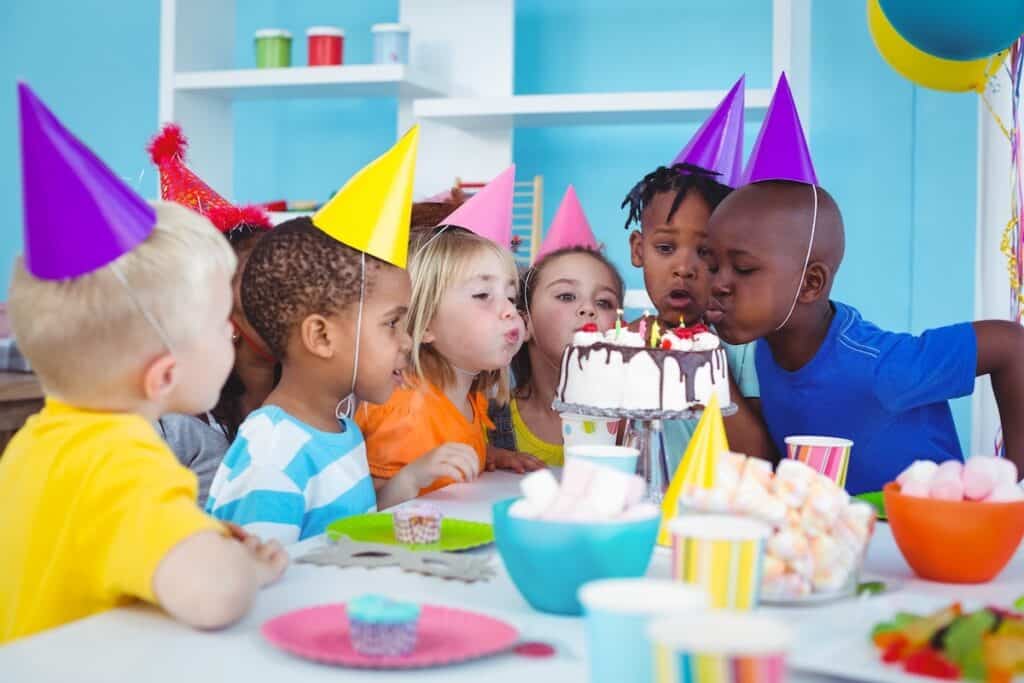 A group of kids wearing party hats at a birthday party sing Happy Birthday and wait to blow out the candles on the cake.