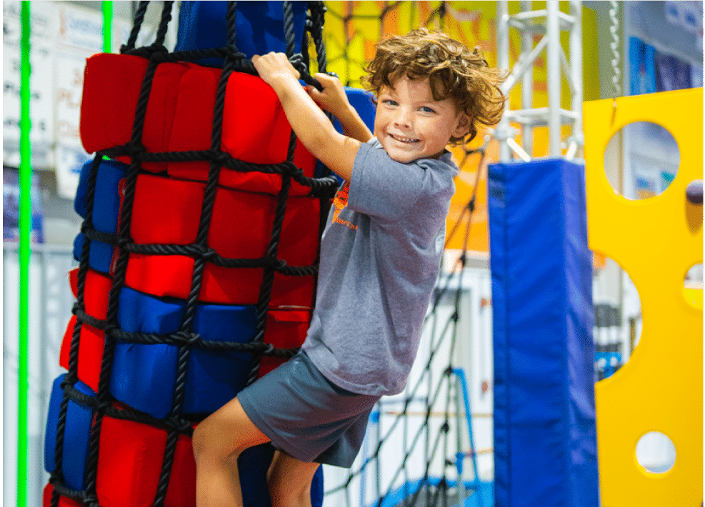 A little boy turns to smile at the camera as he climbs up equipment in the Jungle Gym.