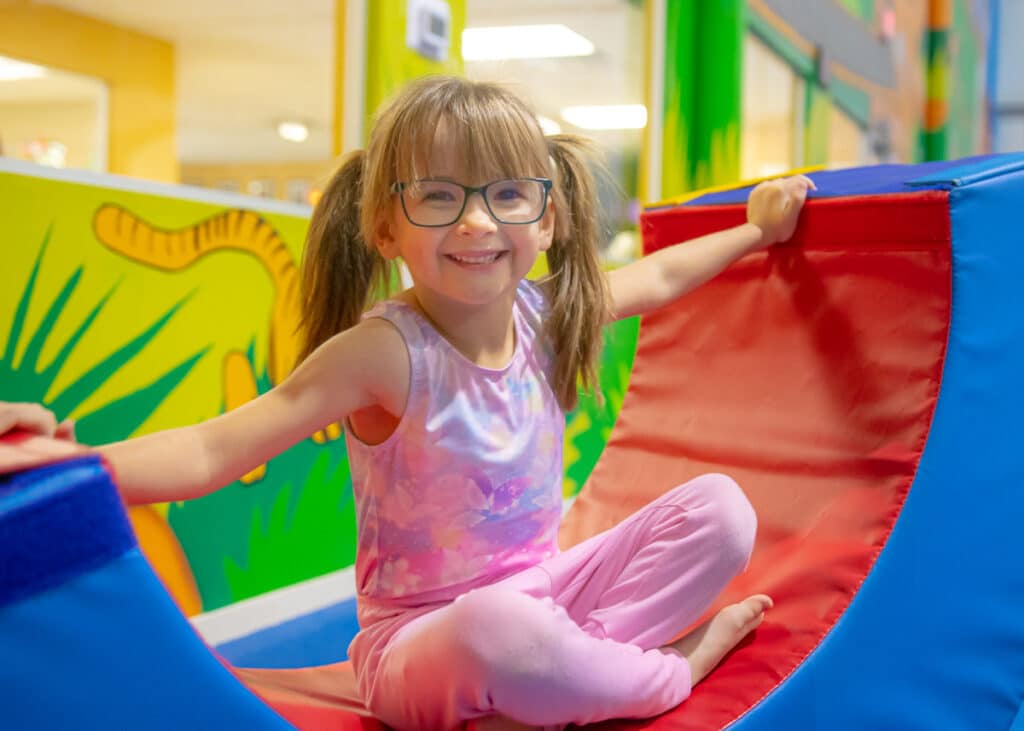 A little girl in glasses and pigtails sits on equipment in the Jungle Gym and smiles.